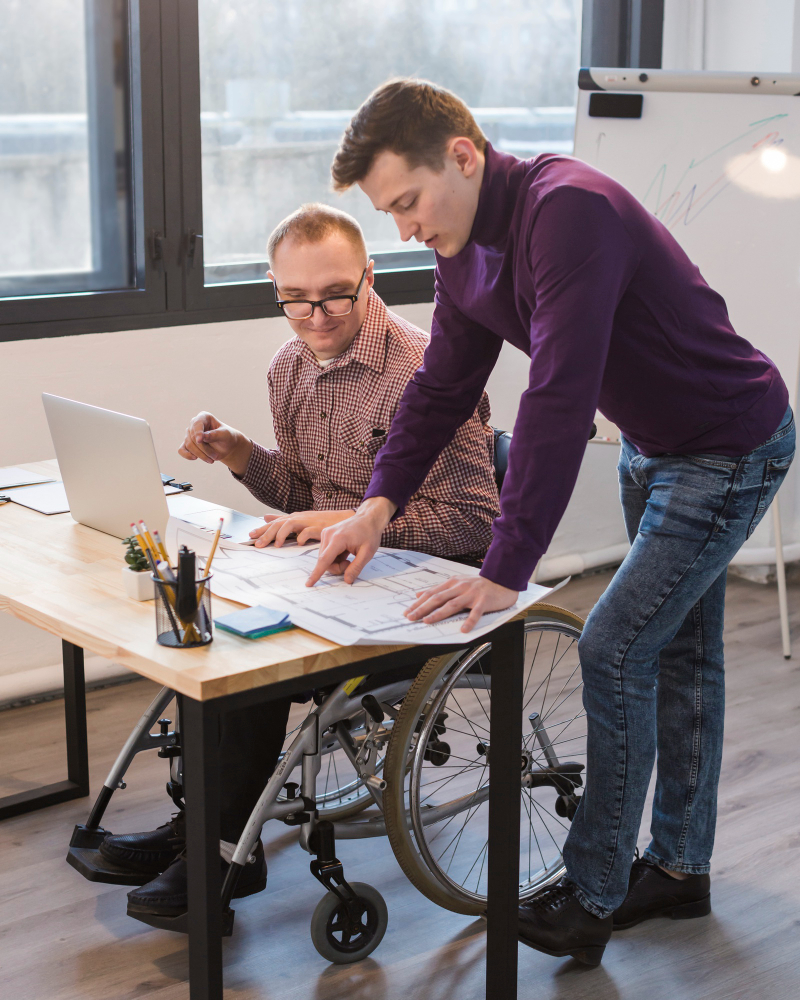 Libera hero header image 2 person working on a desk, one standing points a paper, the other one on a wheelchair and a laptop, looking 
